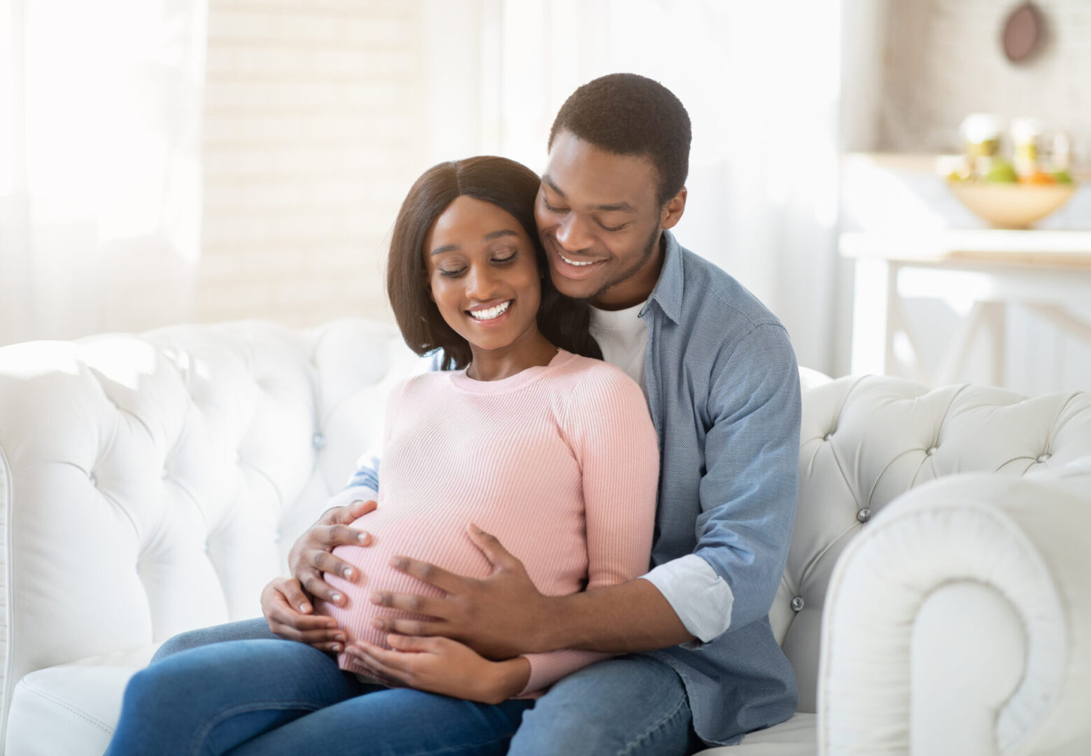 Smiling future mom and dad expecting baby, sitting on sofa at home and hugging. Beautiful pregnant couple embracing on couch. Young African American guy embracing his expectant wife